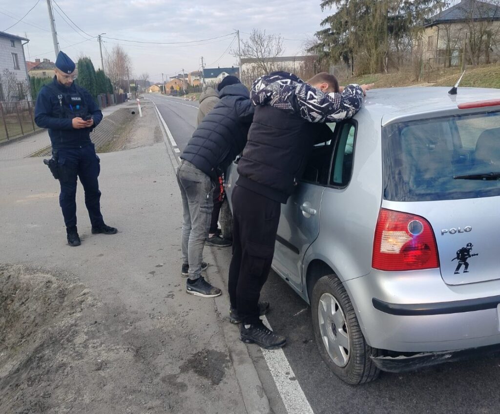 Grupa mężczyzn pochyla się nad otwartymi drzwiami samochodu, podczas gdy policjant stoi obok, przeglądając telefon.