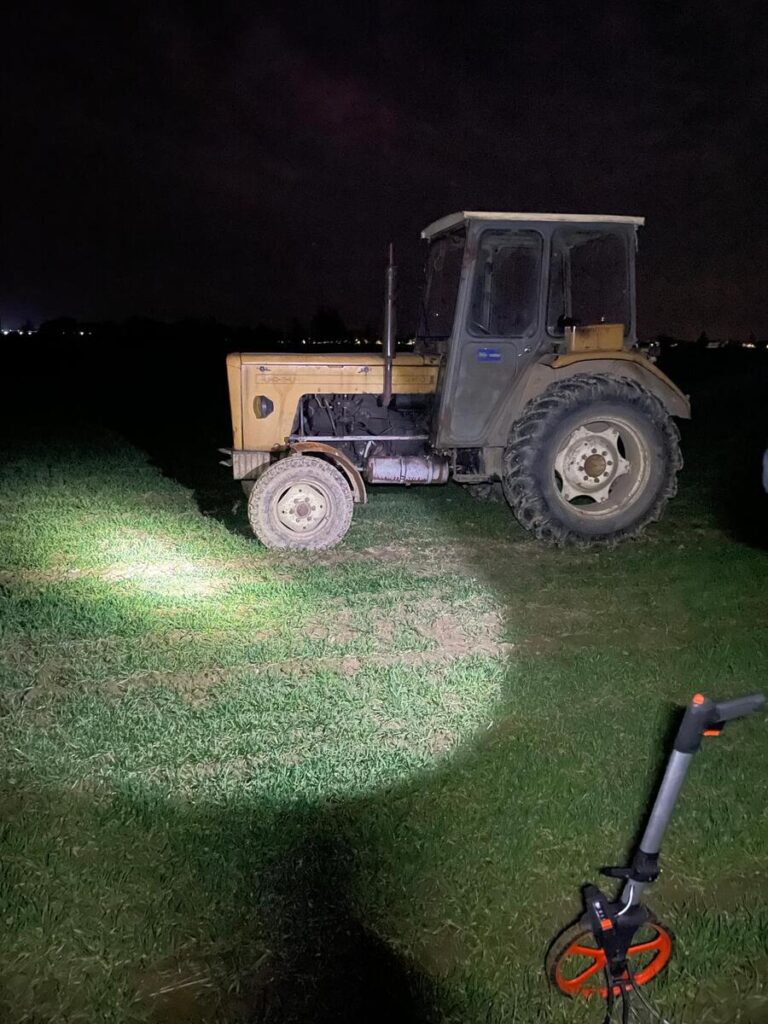 Traktor na polu oświetlony latarką w nocy, w tle widoczne delikatne światła miasta.