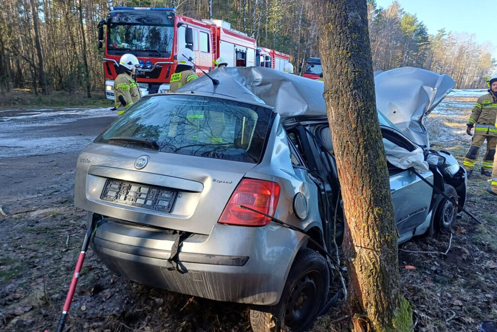 Uszkodzony samochód osobowy zderzony z drzewem na poboczu leśnej drogi, obok strażacy w akcji ratunkowej.