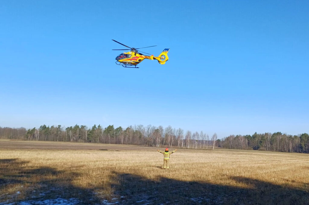 Śmigłowiec ratunkowy unoszący się nad otwartym polem, w tle las, a przed nim stoi mężczyzna w odzieży ochronnej z uniesionymi rękami.
