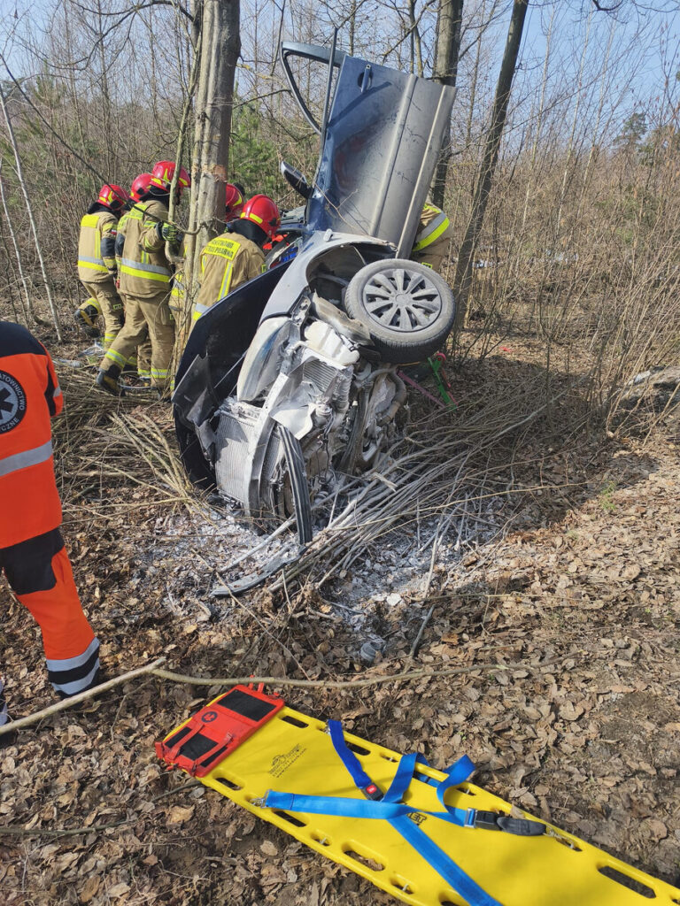 Grupa strażaków w kaskach pracuje nad wydobyciem poszkodowanego z przewróconego samochodu w lesie. Obok leży nosze.
