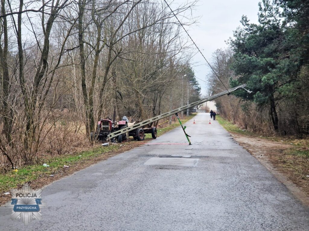 Uszkodzony słup energetyczny leży na drodze, obok ciągnik rolniczy, a w oddali spacerujący ludzie.