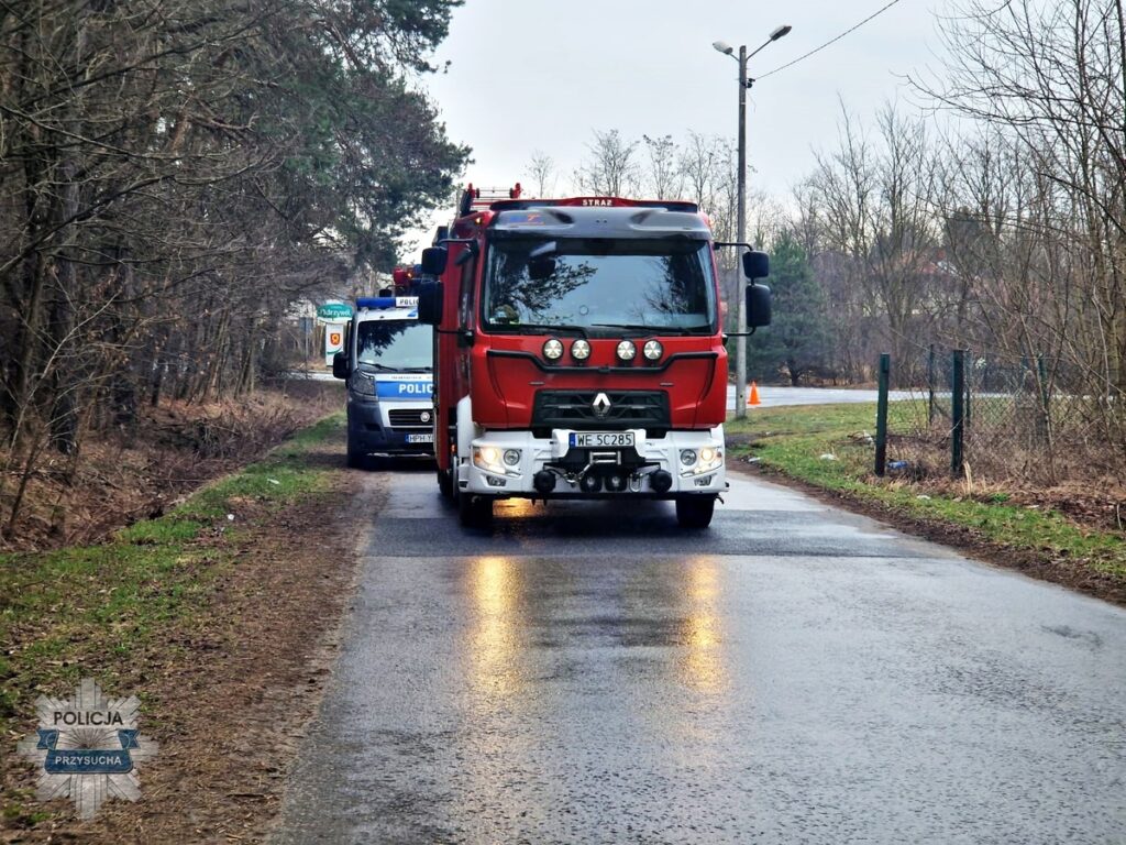 Czerwony wóz strażacki oraz niebieski radiowóz policyjny stoją na wąskiej drodze otoczonej drzewami.