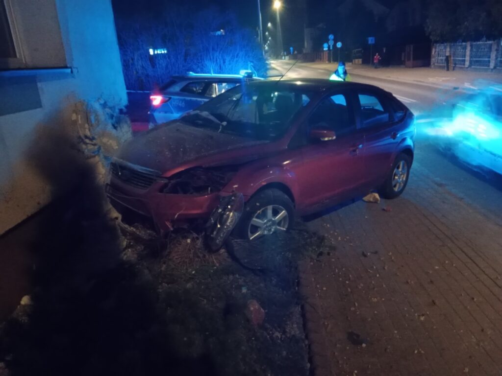 Czerwony samochód osobowy uderzony w ścianę budynku, obok policyjne radiowozy z włączonymi sygnałami świetlnymi.
