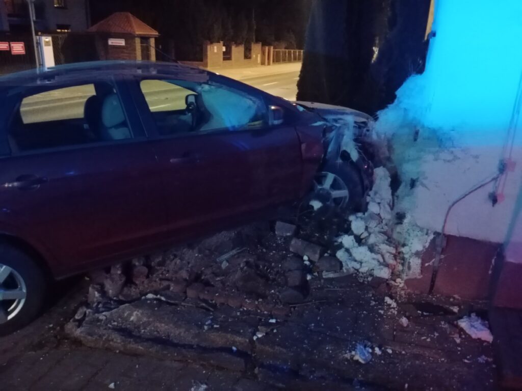 Czerwony samochód osobowy wjechał w ścianę budynku, powodując uszkodzenia i zarysowania na elewacji.
