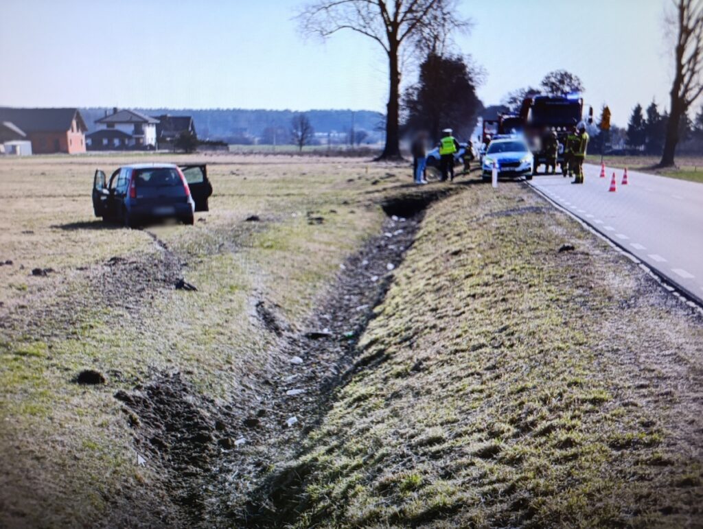 Samochód osobowy stoi na polu z otwartymi drzwiami, obok grupka osób w kamizelkach odblaskowych zabezpiecza miejsce zdarzenia.