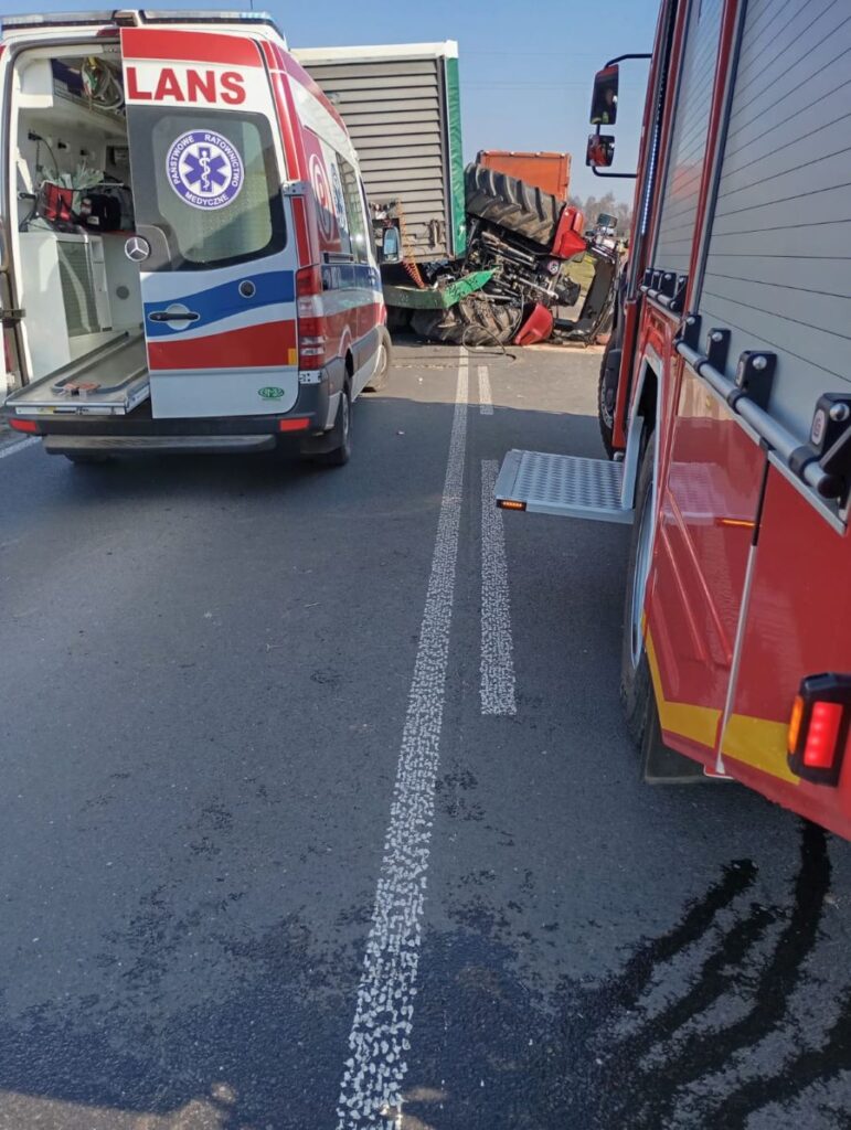 Na drodze leży przewrócony traktor, a obok niego zaparkowane karetka i wóz strażacki.