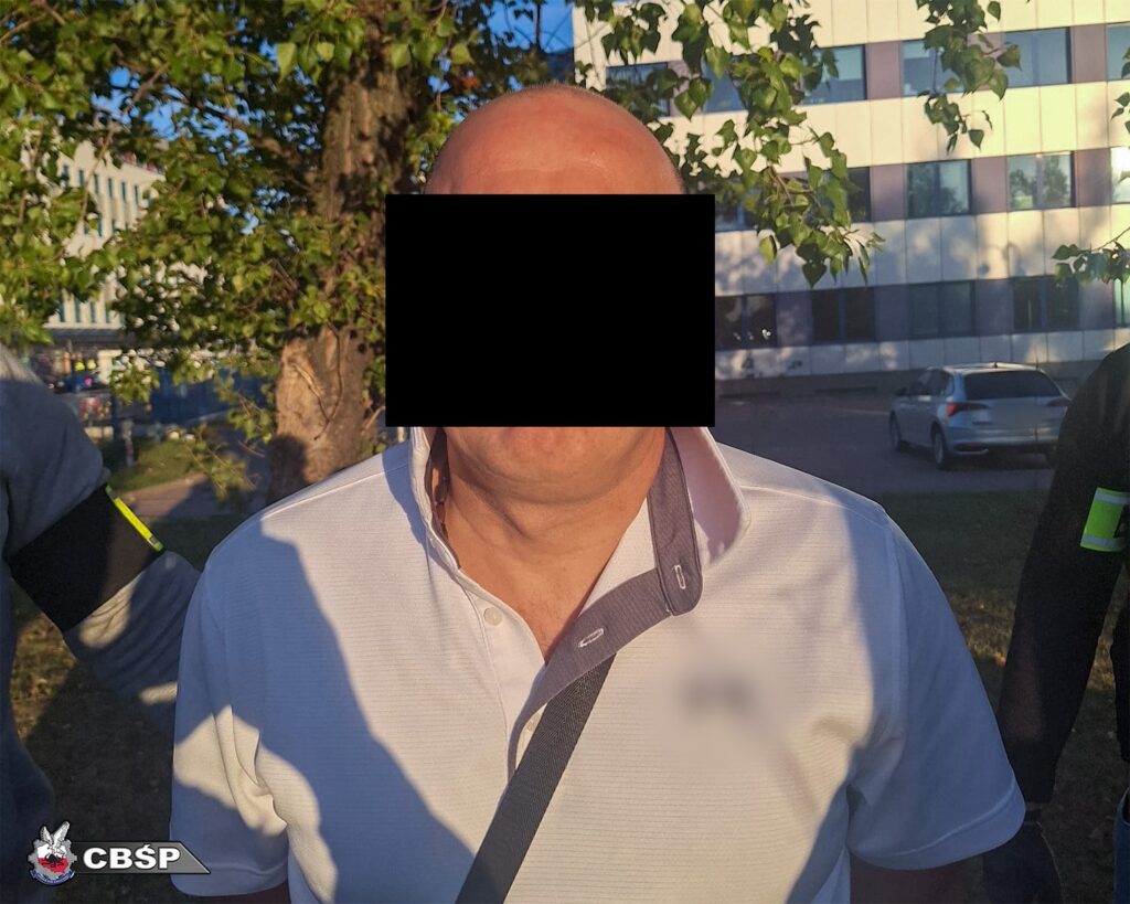 Mężczyzna w białej koszuli stoi w parku, otoczony przez funkcjonariuszy w mundurach. W tle widać nowoczesny budynek.