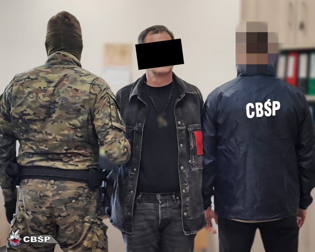 Mężczyzna w czarnej kurtce stoi pomiędzy funkcjonariuszem w mundurze kamuflażowym a osobą w kurtce z logo CBŚP w biurze.