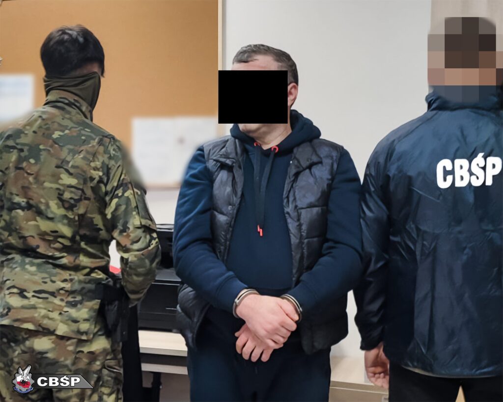 Mężczyzna w dresie stoi z rękami skrępowanymi, obok funkcjonariusz w mundurze z oznaczeniem CBŚP.