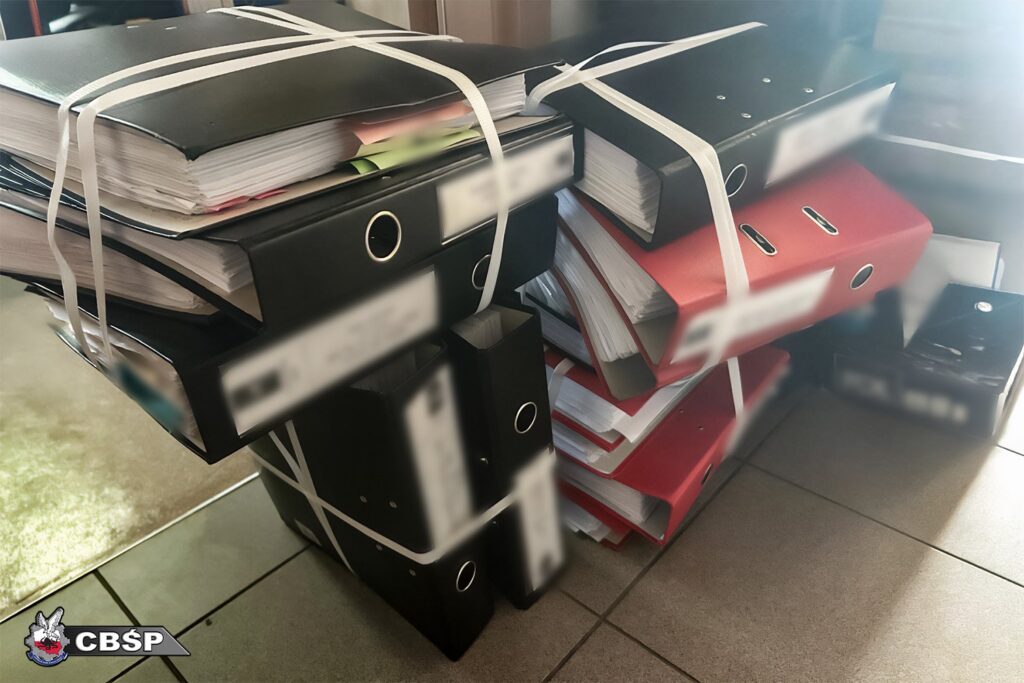 Stosy oprawionych dokumentów w teczkach, związane białymi taśmami, leżą na podłodze w biurze.