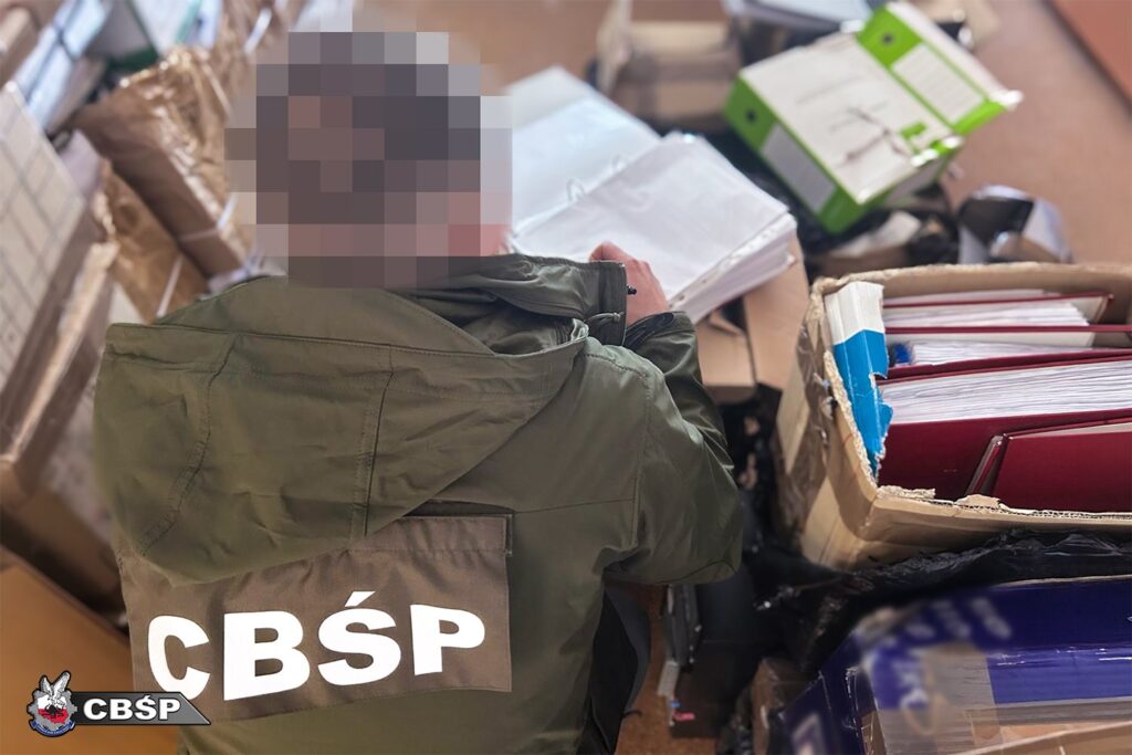 Funkcjonariusz CBŚP przegląda dokumenty w zagraconym pomieszczeniu, otoczony teczkami i segregatorami.