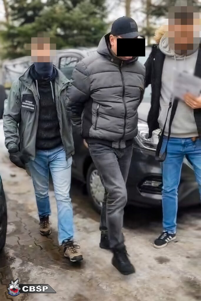 Mężczyzna w szarej kurtce jest prowadzony przez dwóch funkcjonariuszy w kierunku samochodu na parkingu.