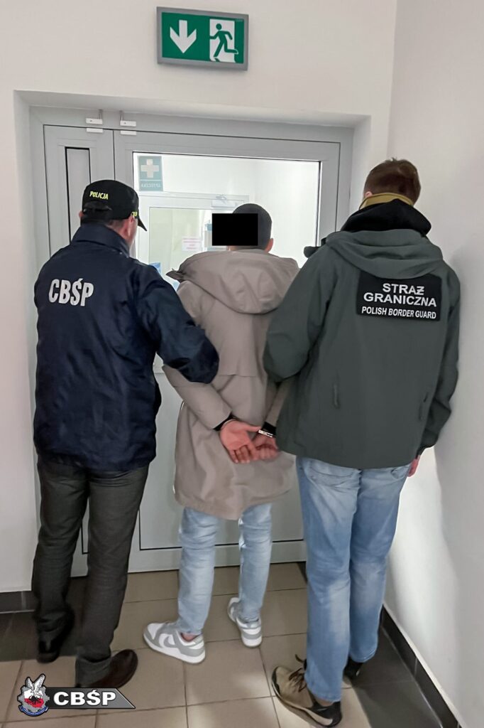 Mężczyzna w kajdankach prowadzony przez funkcjonariuszy CBŚP i Straży Granicznej w budynku z jasnymi ścianami.