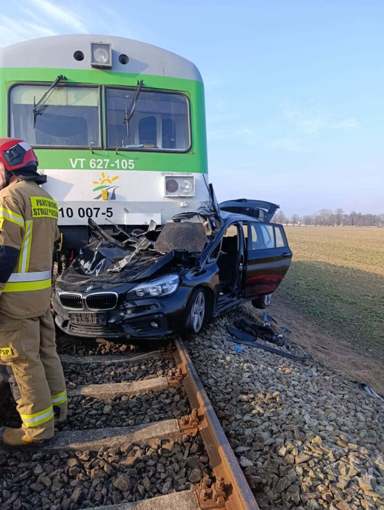 Czarny samochód osobowy uszkodzony po zderzeniu z pociągiem na torach, obok strażak w mundurze interweniujący na miejscu zdarzenia.