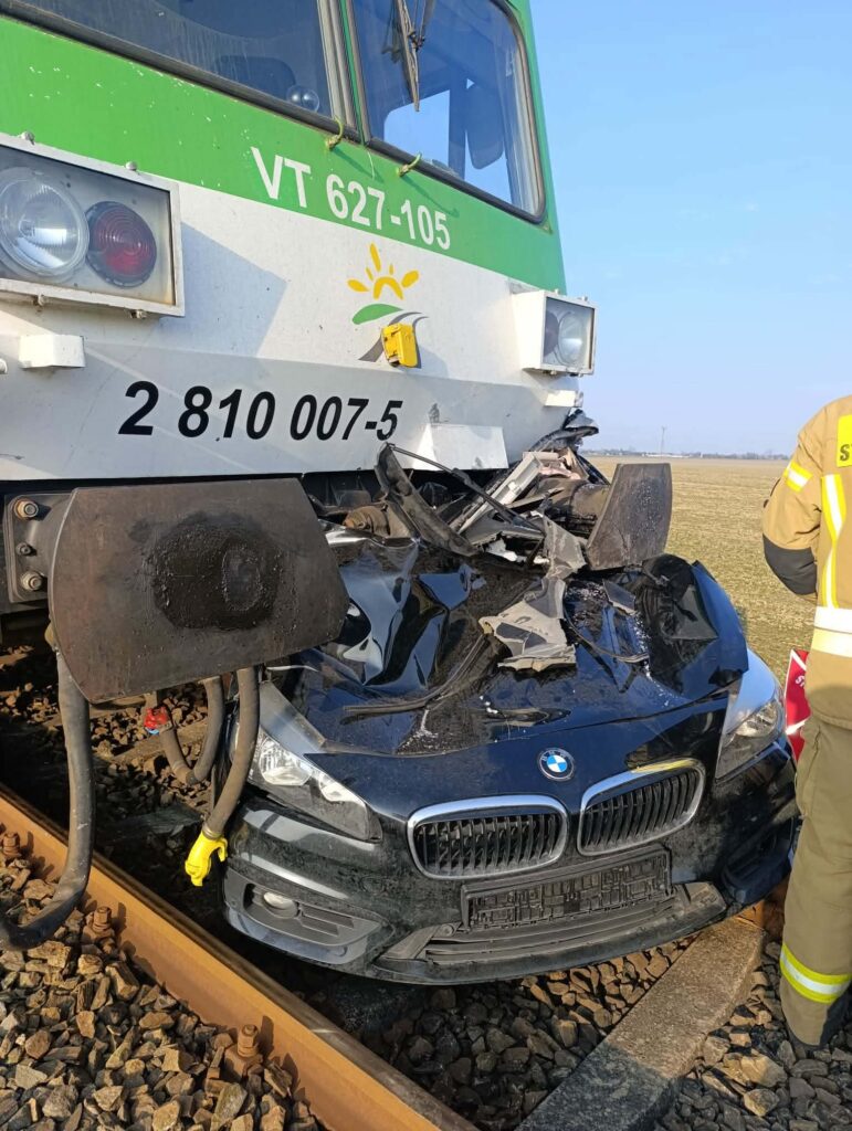 Pociąg uderzył w rozbity czarny samochód osobowy na torach w otwartym terenie. Obok stoi strażak.