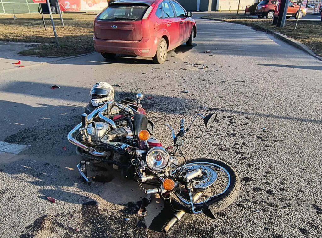 Motocykl leżący na drodze z uszkodzeniami, obok stoi czerwony samochód, na jezdni widać rozbite szkło.
