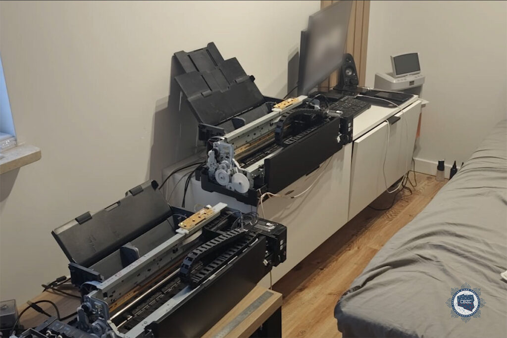 Dwa rozmontowane urządzenia drukarskie stoją na białej szafce w jasnym pomieszczeniu, obok łóżka i monitora.