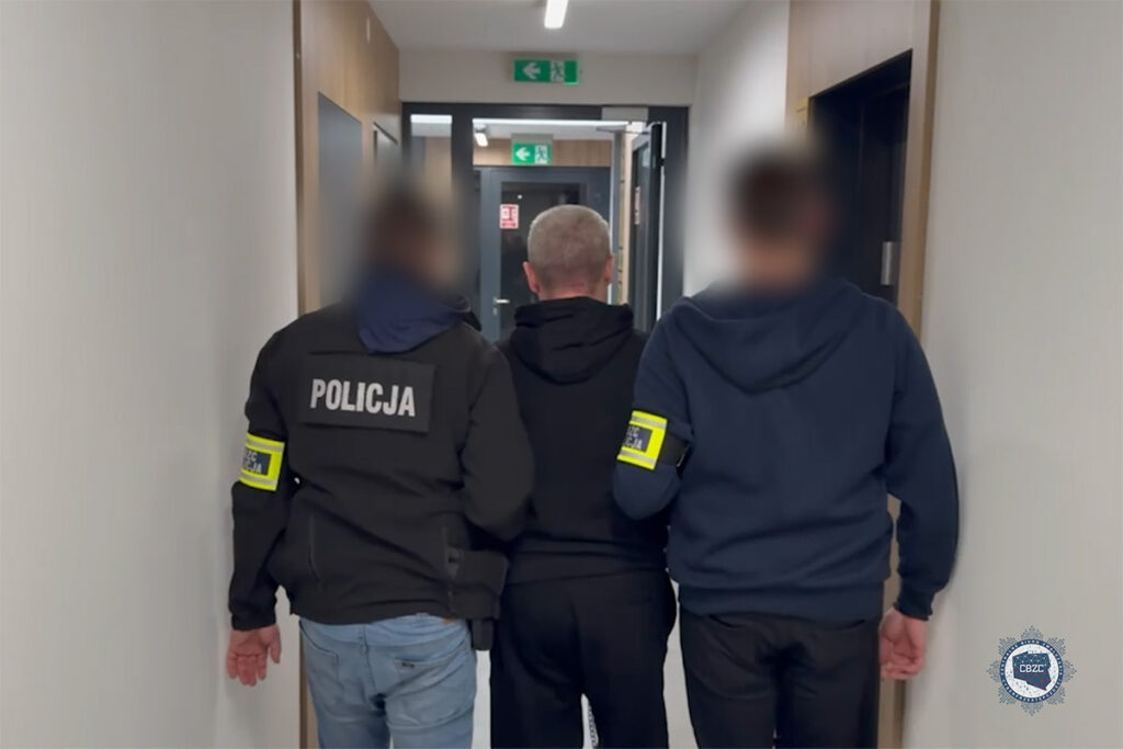 Policjanci prowadzą podejrzanego korytarzem w budynku, w tle drzwi i znaki ewakuacyjne.