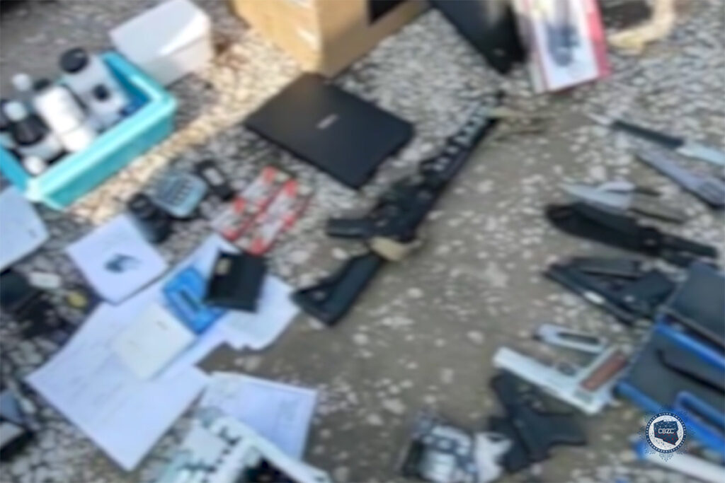 Na ziemi leżą różne przedmioty, w tym broń palna, dokumenty oraz sprzęt elektroniczny, w nieuporządkowanej aranżacji.