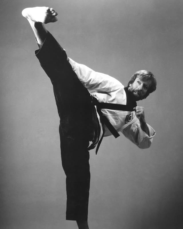 Mężczyzna w judodze wykonujący wysokie kopnięcie w czarnym pasie na tle szaro-białym.