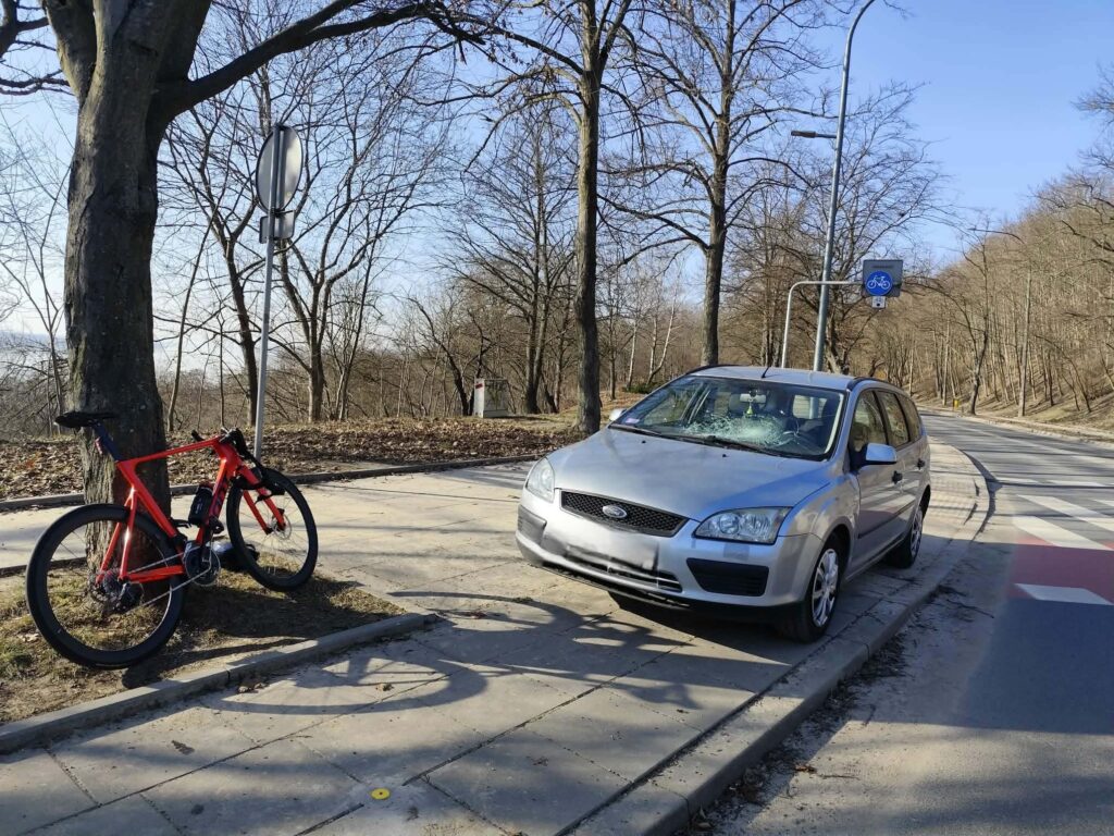 Czerwony rower stoi przy drzewie obok zaparkowanego srebrnego samochodu na wybrukowanej ścieżce.