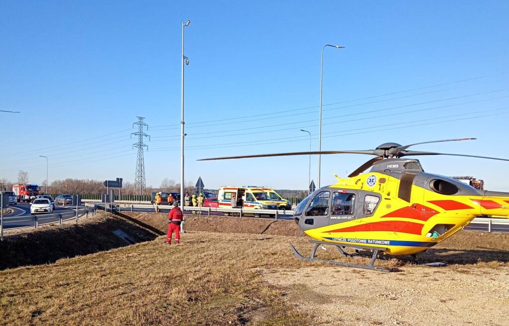 Na poboczu drogi lądował śmigłowiec ratunkowy, obok pracują ratownicy medyczni. W tle widać samochody i niebieskie niebo.
