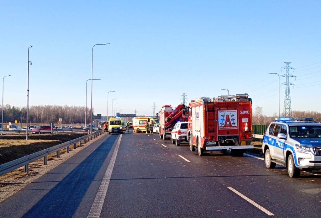 Na drodze stoi zespół ratunkowy, policyjny radiowóz oraz wóz strażacki, a w tle widać niebieskie niebo.