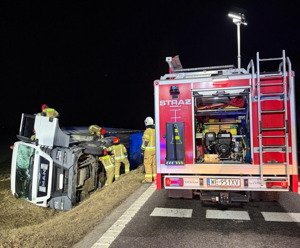 Strażacy w żółtych mundurach zabezpieczają przewrócony samochód ciężarowy na poboczu drogi w nocy.