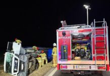 Strażacy w żółtych mundurach zabezpieczają przewrócony samochód ciężarowy na poboczu drogi w nocy.