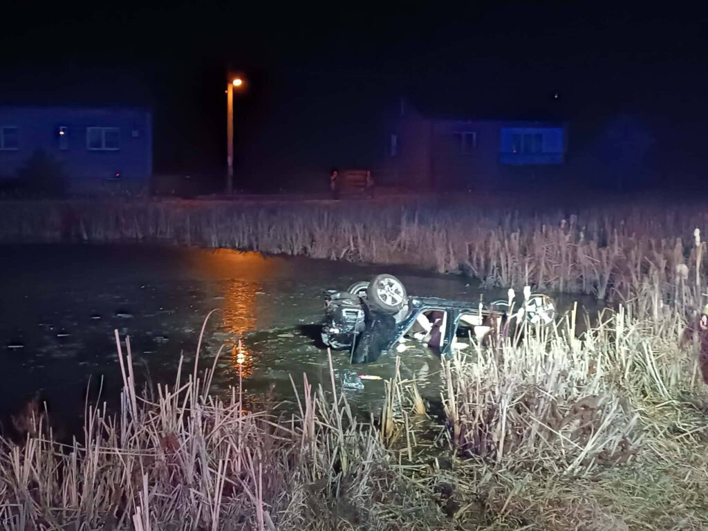 Samochód leży na dachu w wodzie, otoczony przez trzciny, w tle widać oświetlony budynek i latarnię.