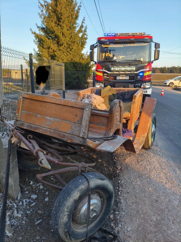 Zniszczony wóz z drewnianą konstrukcją leży przy poboczu drogi, obok strażackiego wozu z włączonymi sygnałami świetlnymi.