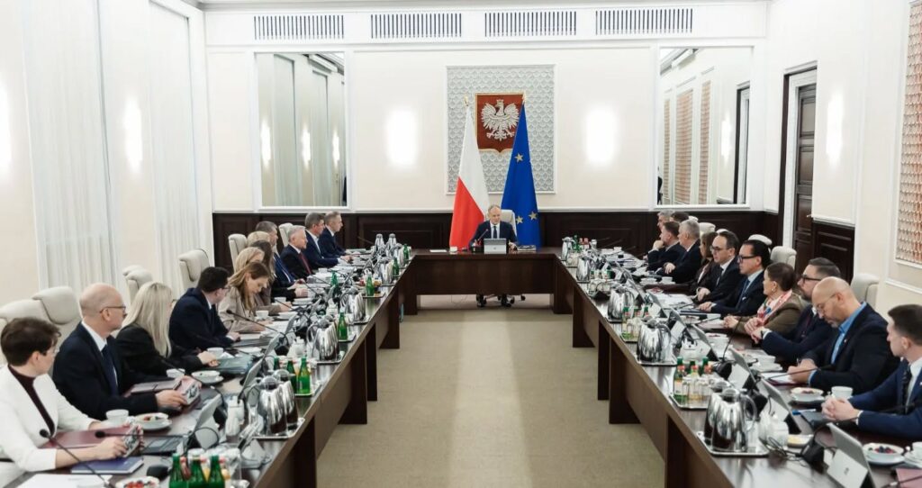 Członkowie rządu siedzą przy długim stole w nowoczesnej sali konferencyjnej, prowadząc dyskusję.