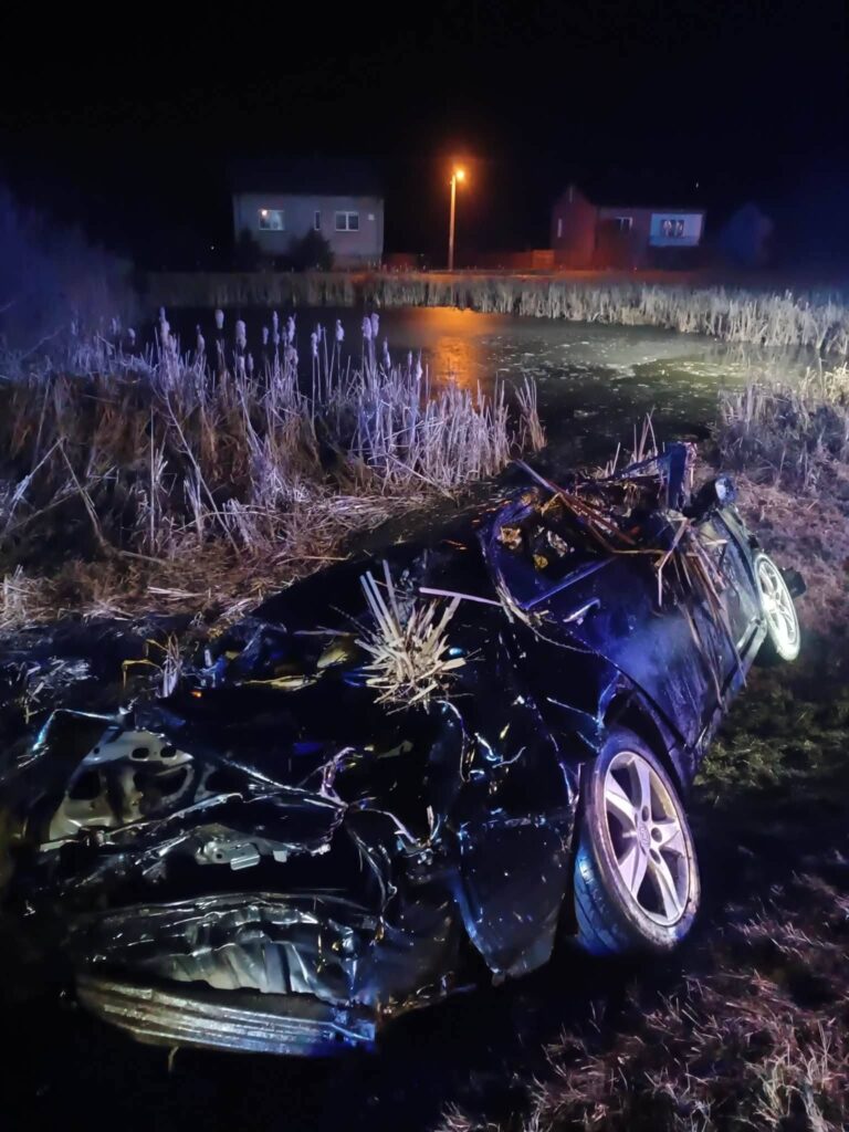 Zniszczony samochód leży na boku w pobliżu stawu, otoczony przez trawę i oświetlenie uliczne w nocy.
