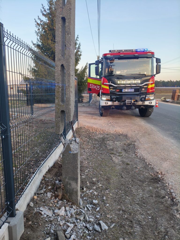 Straż pożarna z włączonymi sygnałami świetlnymi stoi obok uszkodzonego słupa betonowego przy drodze.