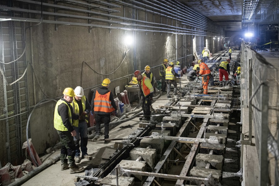 Grupa pracowników budowlanych w kaskach i kamizelkach odblaskowych wykonuje prace w podziemnym tunelu.