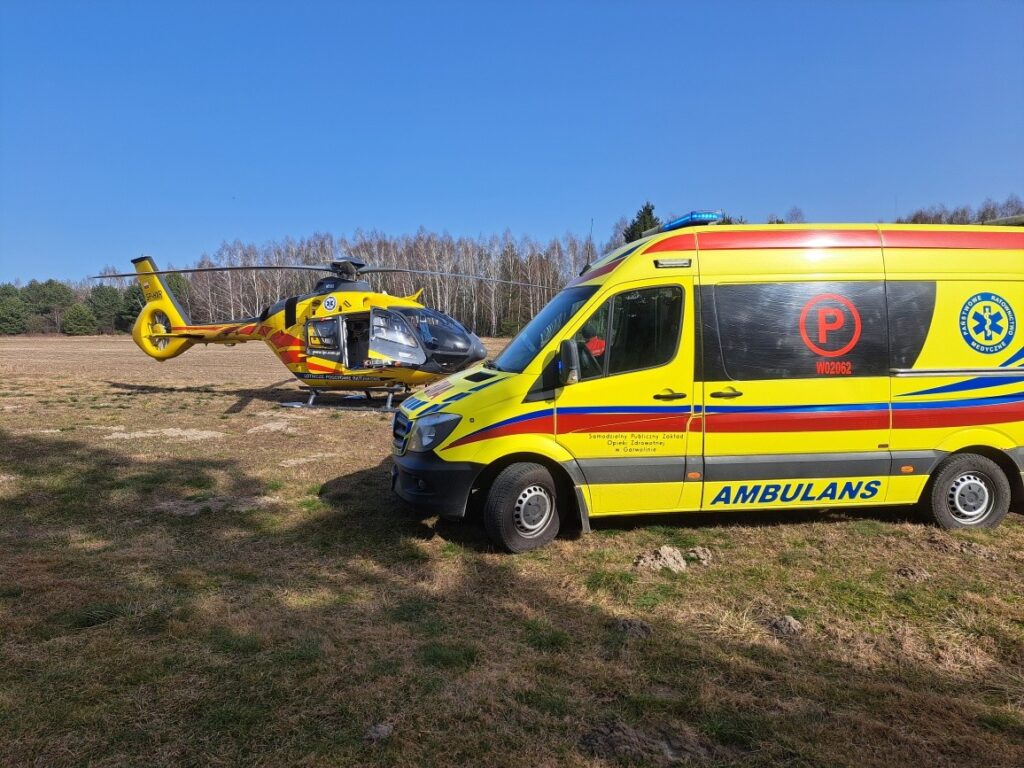 Śmigłowiec ratunkowy i ambulans stoją na otwartej przestrzeni, otoczonej lasem, w słoneczny dzień.