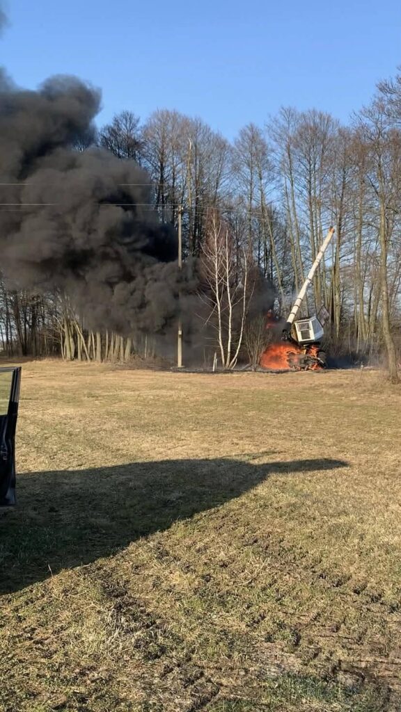 Maszyna leśna płonąca w lesie, w tle czarny dym unoszący się w niebo, otoczenie z drzewami i trawą.