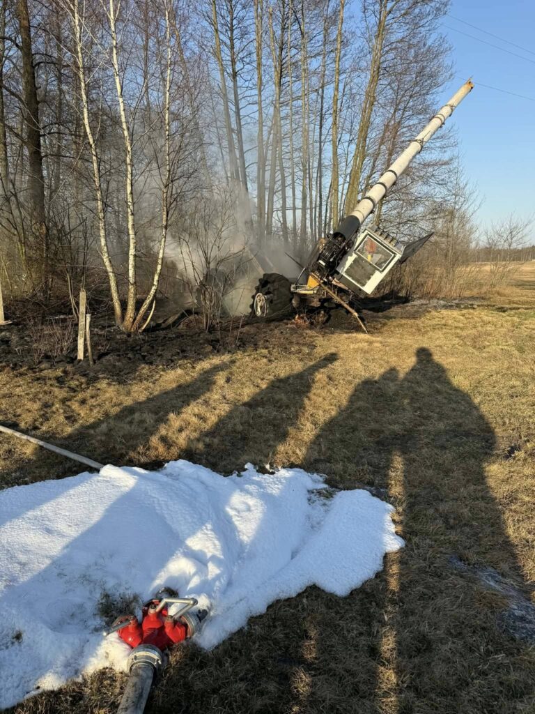 Maszyna budowlana przewrócona w lesie, w tle widoczne zarośla i dym, w pierwszym planie wąż strażacki z pianą.