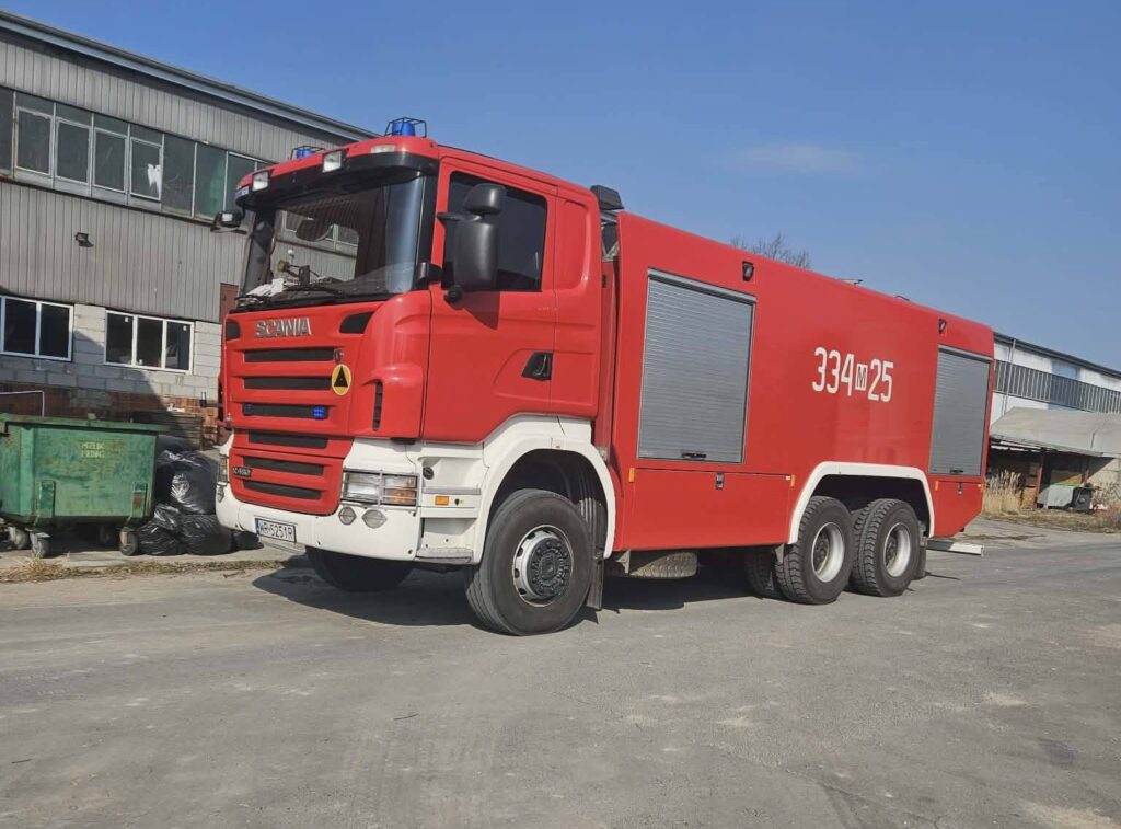 Czerwony wóz strażacki marki Scania zaparkowany na terenie przemysłowym obok budynków i kontenerów.