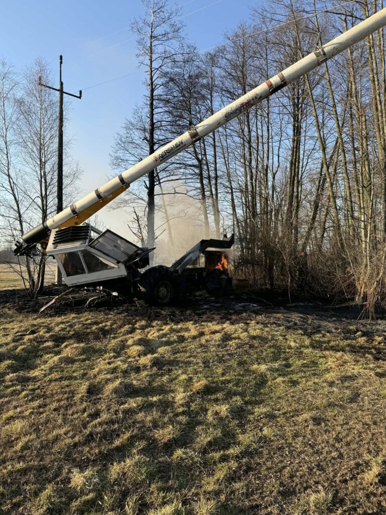 Maszyna budowlana z uniesioną wysięgnikiem stoi w pobliżu zarośli, w tle widać dym i ogień.