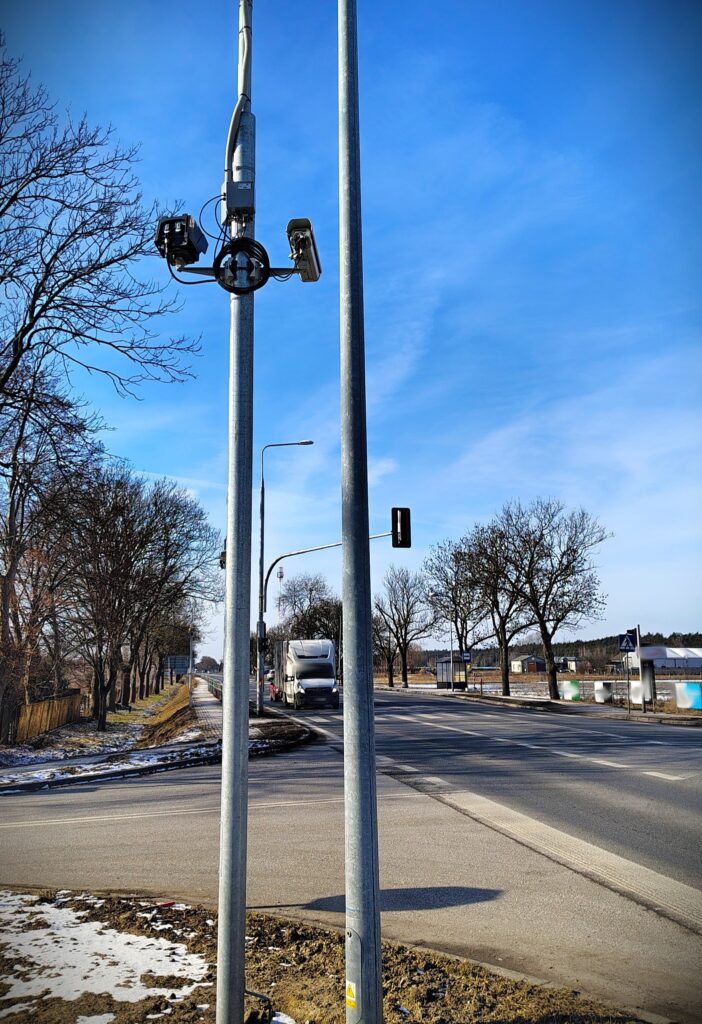 Kamery monitorujące zamontowane na słupie, obok drogi z widokiem na przejeżdżający ciężarówkę i drzewa w tle.