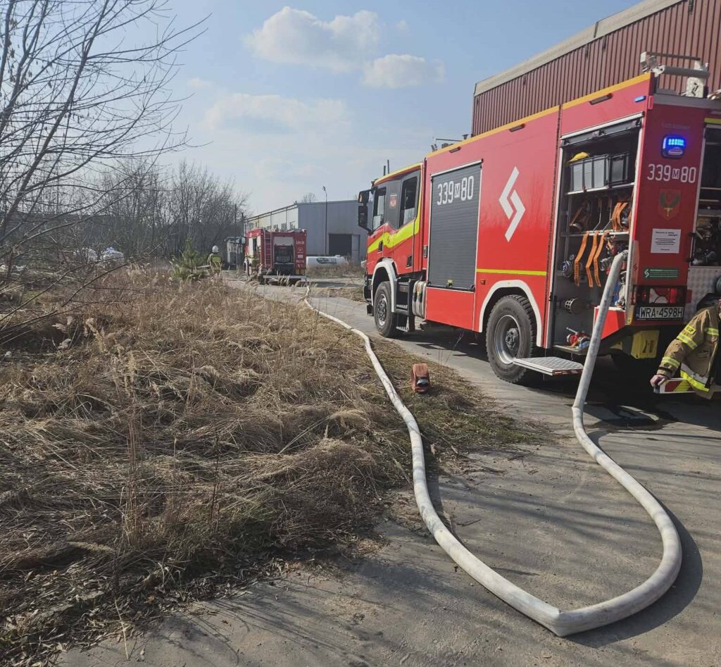 Czerwony wóz strażacki z rozwiniętym wężem na terenie przemysłowym, w otoczeniu wysokiej trawy i budynków.