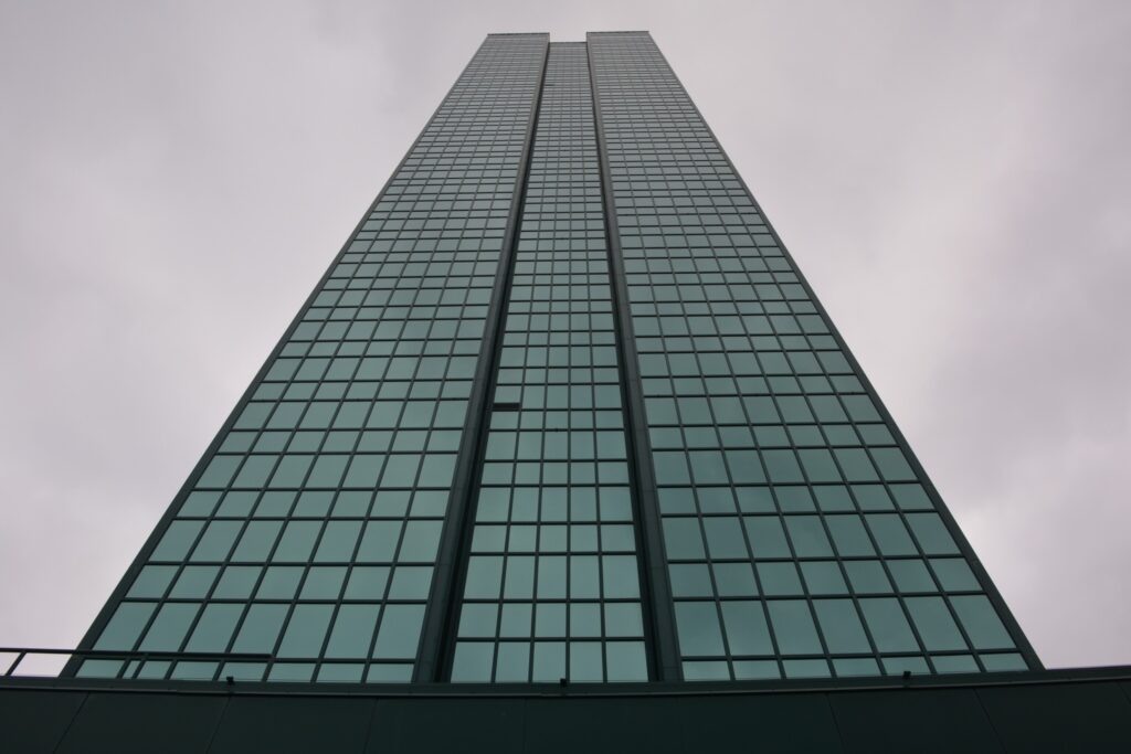 Wysoki biurowiec z przeszkloną fasadą, widoczny z dołu, na tle pochmurnego nieba.