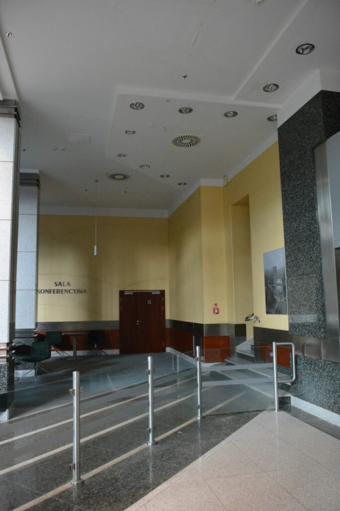 Przestronna sala konferencyjna z jasnymi ścianami, eleganckim oświetleniem oraz szklanymi barierkami.