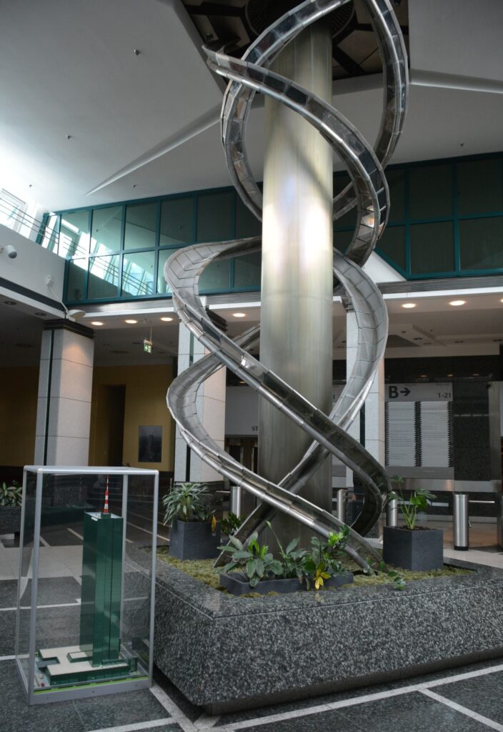 Metalowa spirala umieszczona w nowoczesnym wnętrzu biurowca, obok modelu zielonego wieżowca. Zielone rośliny zdobią przestrzeń.