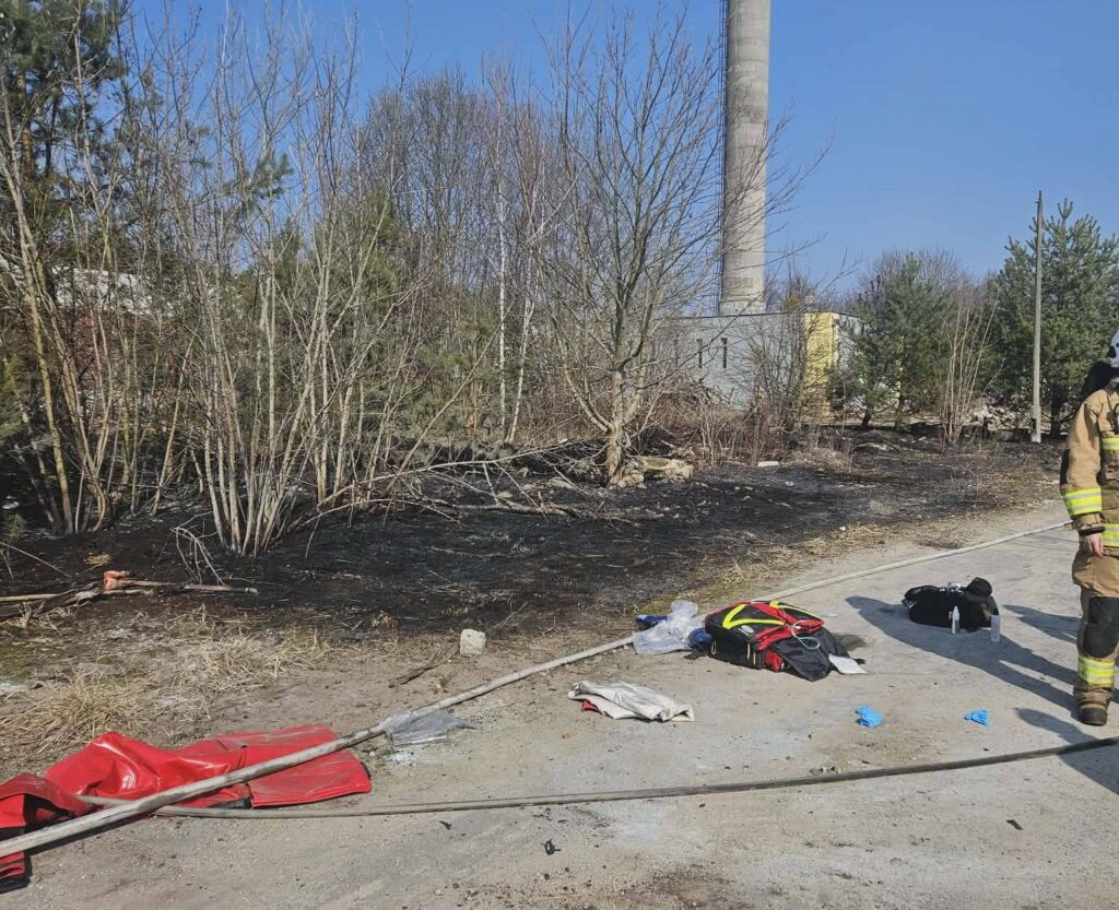 Strażak w mundurze monitoruje teren po pożarze, na którym widać spalone krzewy i leżące torby ratunkowe.