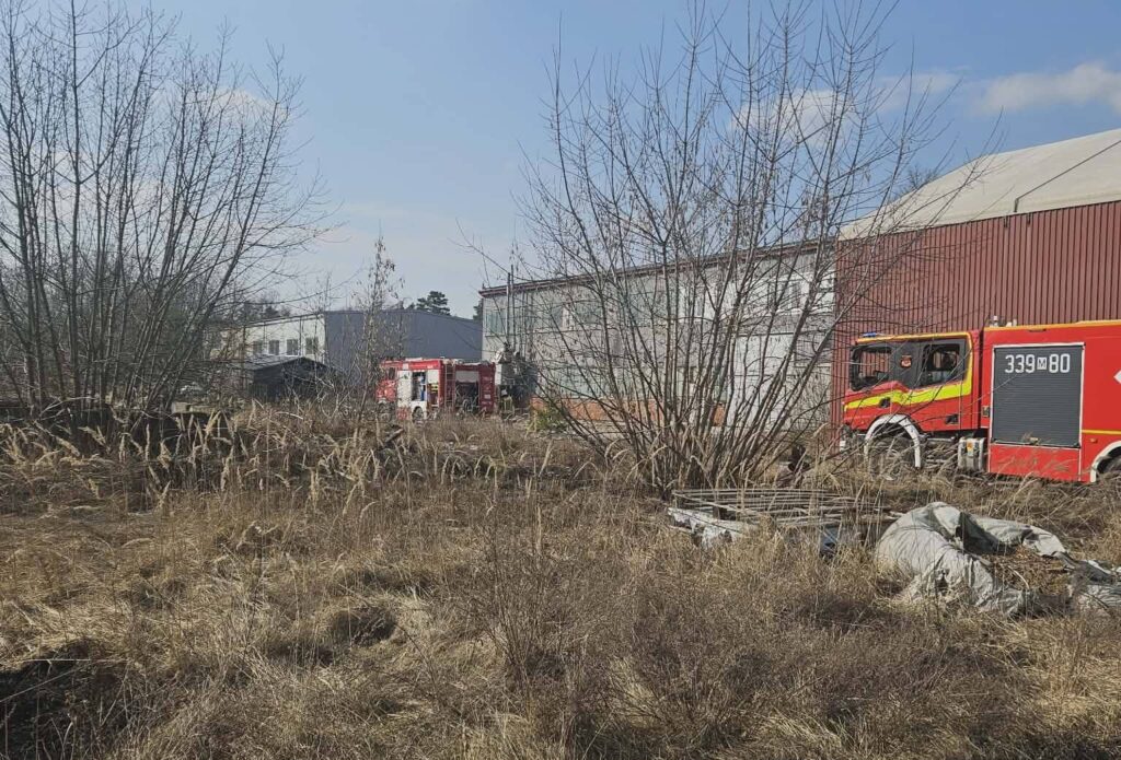 W tle zarośnięte tereny, widać wóz strażacki zaparkowany obok budynków przemysłowych.