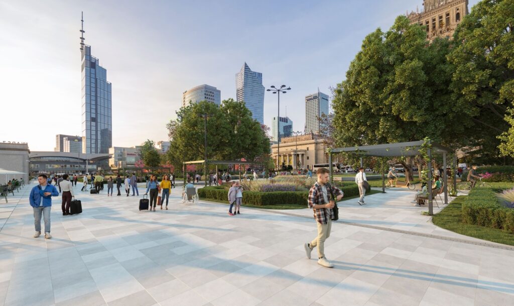 Grupa ludzi spaceruje po nowoczesnym placu z zielenią, w tle widać wieżowce i budynki biurowe.
