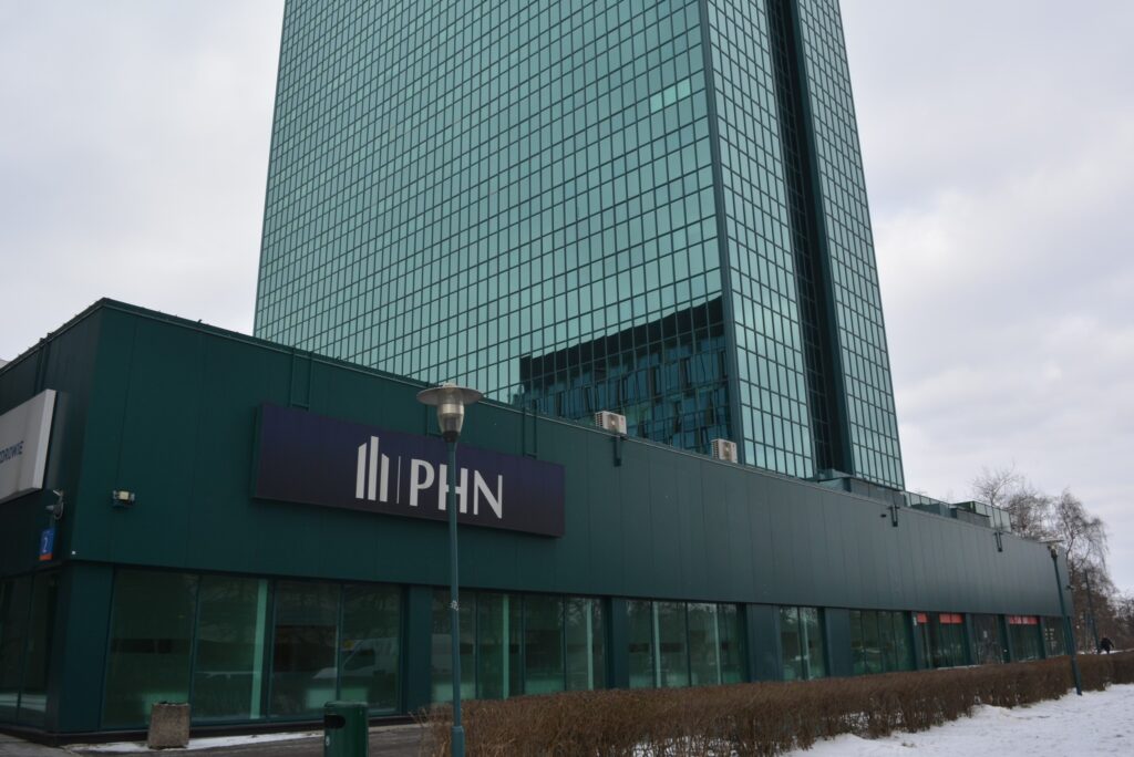 Nowoczesny biurowiec z przeszkloną fasadą i dużym logo "PHN" na zielonej elewacji, otoczony niskim żywopłotem.